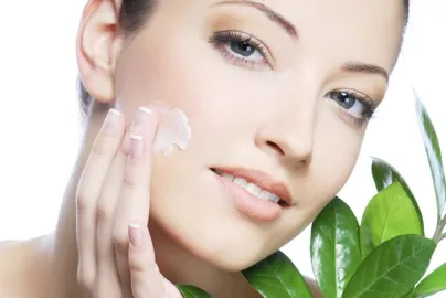A woman smoothing cream over her cheek, with green leaves beside her